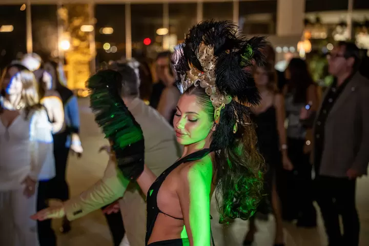 Performer con copricapo di piume nere e ventaglio tra gli ospiti, in luce verde