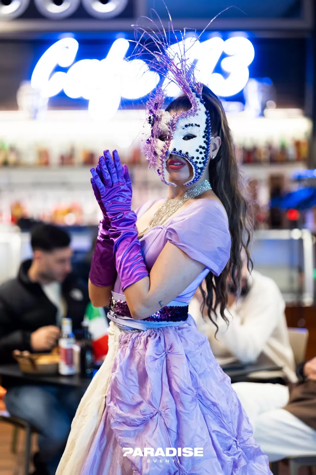 Performer con maschera decorata, abito lilla e guanti viola tra i tavoli di un locale